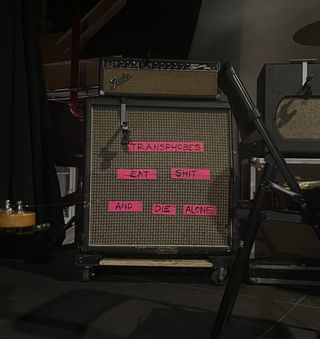 "A bass amplifier on stage, surrounded by some other gear. Pink tape runs across the surface. Written on this are the words «Transphobes eat shit and die alone."