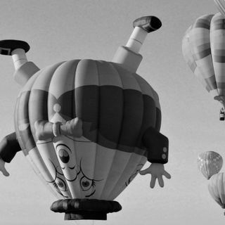 Some hot air baloons drifting through the sky. The one in the foreground is in the form of an upside down slightly uncomfortable looking cartoon character