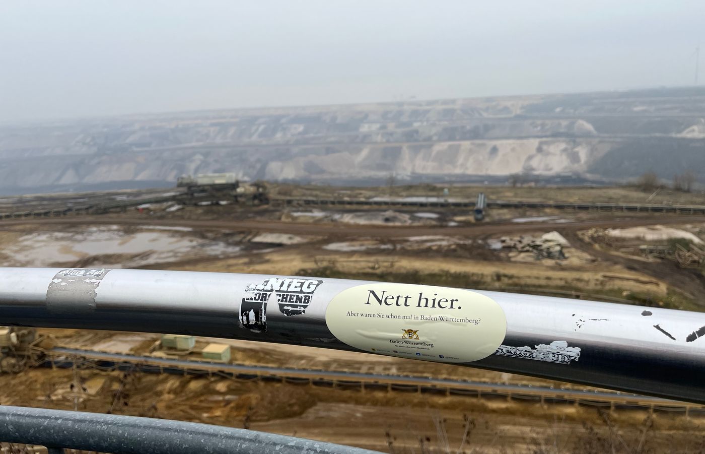 "View into the coal mine Garzweiler. In the front you can see a guard rail on which someone put a sticker reading «Nett hier». The background is blurry and shows the mine itself. Everything is brown, all shades of brown. The sky is cloudy and the view partially obscured by layers of fog."