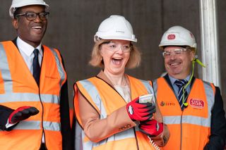 A picture by Liz Truss. She is wearing a safety helmet and a high-visibility waistcoat. In her hand she is holding a button. She looks slightly insane. To her left is Kwasi Kwarteng. To her right is an elderly man.